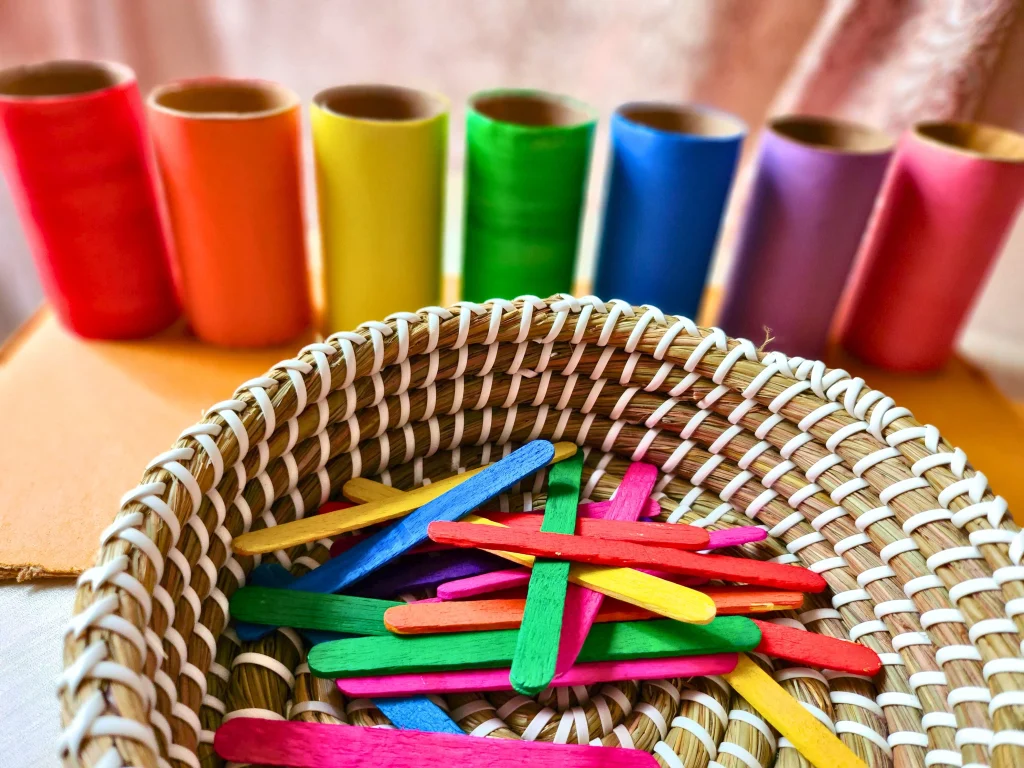 a basket half filled with colorful popsicle sticks and colored toilet paper rolls array behind it