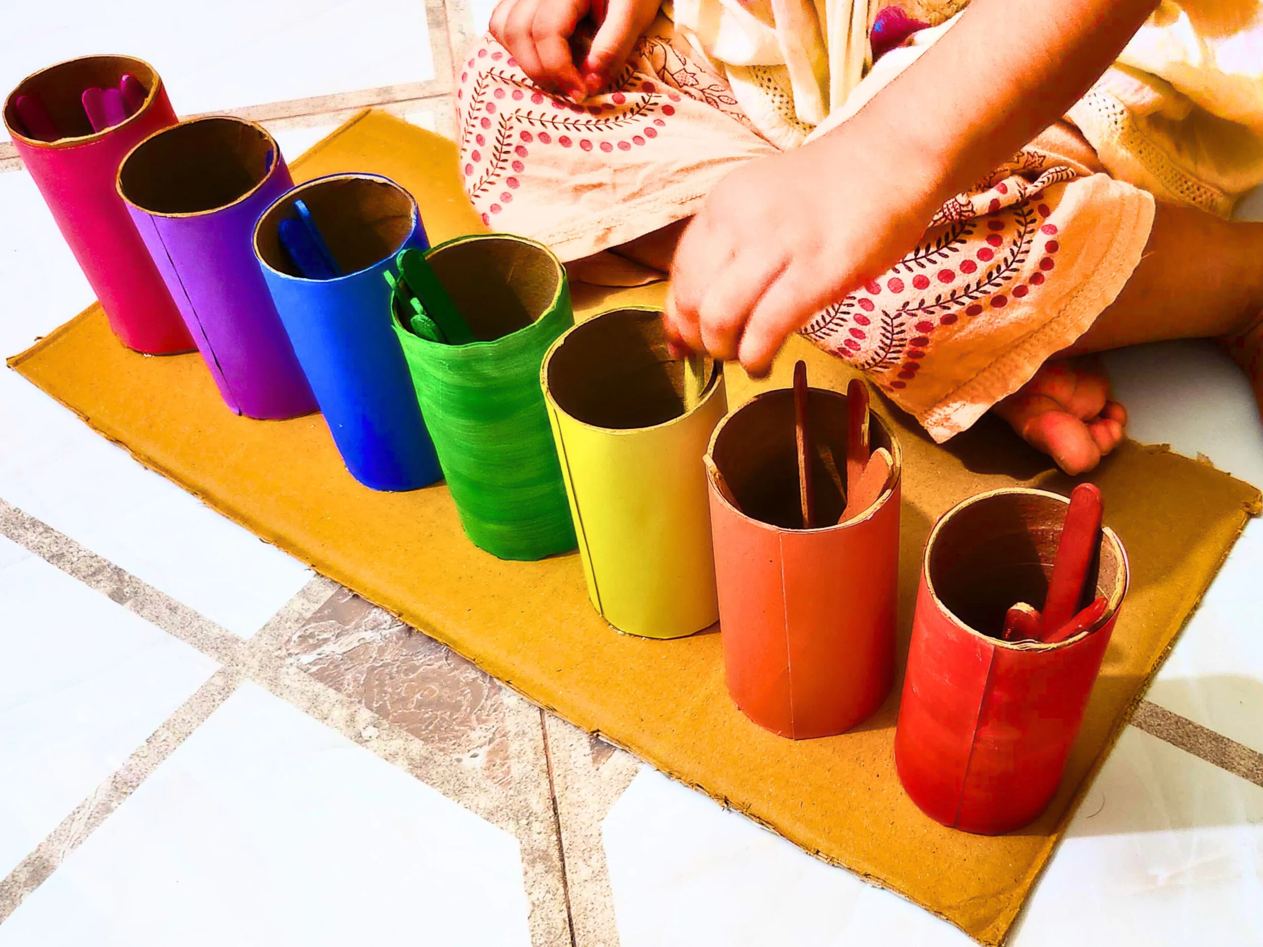 a toddler is sorting colors into DIY colored toilet paper rolls using colored popsicle sticks 